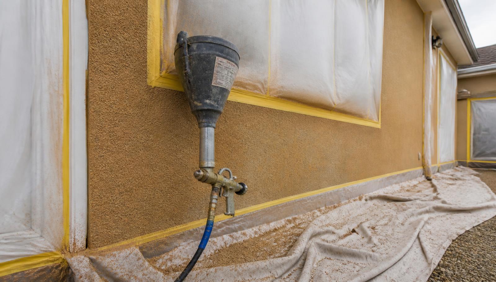 A hopper spray gun and compressor hose against a masked house wall